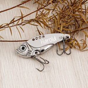May include: A silver-colored fishing lure shaped like a fish, with the name "Harold" engraved on its side. The lure has a weight of 15g and features two treble hooks. The background includes dried brown foliage on a light wooden surface.
