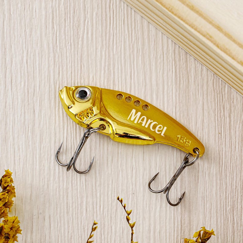 May include: A gold-colored fishing lure shaped like a fish, with the name "Marcel" printed on its side. The lure has two treble hooks and the weight is 15g. The lure is on a light-colored wooden surface.