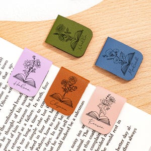 Peut inclure: Cinq marque-pages en cuir de couleurs variées : vert, bleu, marron, rose et lavande. Chaque marque-page présente un motif floral au-dessus d'une illustration de livre ouvert, avec un nom en dessous. Les marque-pages sont sur une surface en bois.