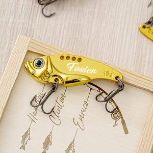 May include: A gold-colored fishing lure with the name "Fosten" and the weight "15g" printed on it. The lure has a fish-like shape with a hook and eye detail. The lure is displayed in a wooden frame.