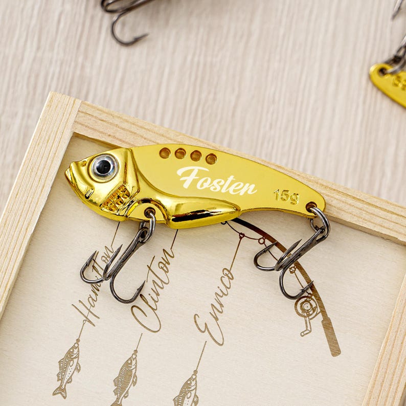 May include: A gold-colored fishing lure with the name "Fosten" and the weight "15g" printed on it. The lure has a fish-like shape with a hook and eye detail. The lure is displayed in a wooden frame.