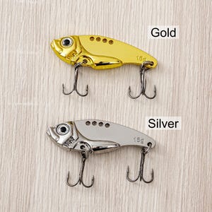May include: Two fishing lures, one gold and one silver, shaped like fish. Each lure has two treble hooks and the text "15g". The word "Gold" is above the gold lure, and "Silver" is above the silver lure.