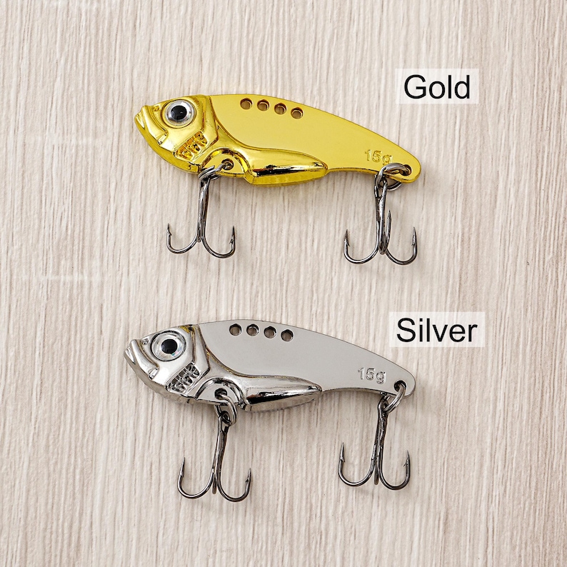 May include: Two fishing lures, one gold and one silver, shaped like fish. Each lure has two treble hooks and the text "15g". The word "Gold" is above the gold lure, and "Silver" is above the silver lure.