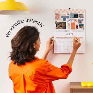 Puede incluir: Un calendario de pared con el mes de julio, adornado con fotos y pegatinas. La persona est&aacute; personalizando el calendario con un bol&iacute;grafo. Las palabras "Personalise Instantly" est&aacute;n escritas en la pared. La persona lleva una camisa naranja.