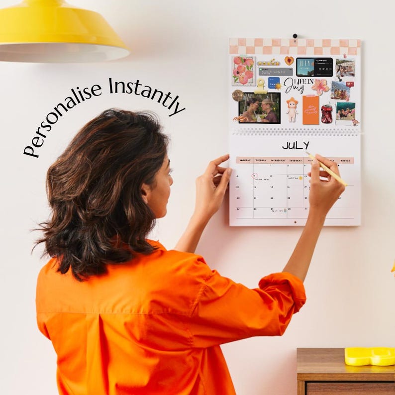 Puede incluir: Un calendario de pared con el mes de julio, adornado con fotos y pegatinas. La persona est&aacute; personalizando el calendario con un bol&iacute;grafo. Las palabras "Personalise Instantly" est&aacute;n escritas en la pared. La persona lleva una camisa naranja.