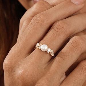 Puede incluir: Un anillo dorado con una perla blanca redonda en el centro, flanqueada por piedras preciosas transparentes más pequeñas. El anillo se muestra en una mano, destacando el diseño intrincado. Una joya elegante.