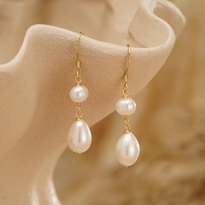 May include: A pair of elegant gold-tone drop earrings. Each earring features a small, round pearl suspended above a larger, teardrop-shaped pearl. The earrings are displayed against a neutral, beige background, highlighting their delicate design and luster.