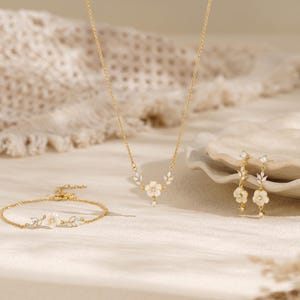 Peut inclure: Un ensemble de bijoux doré composé d'un collier, d'un bracelet et d'une paire de boucles d'oreilles pendantes. Chaque pièce présente un motif de fleur blanche centrale orné de petits cristaux clairs et de feuilles dorées délicates.