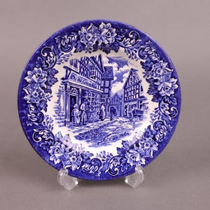 Pode incluir: Prato de cerâmica azul e branco com uma ilustração detalhada de uma pitoresca cena de uma aldeia europeia. O prato apresenta um edifício com uma placa que diz "The Old Curiosity Shop" e é rodeado por intrincados desenhos florais.