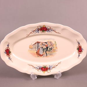 Puede incluir: Un plato ovalado blanco para servir con un diseño floral y una escena de personas bailando en un pueblo. El plato tiene un borde elevado y un borde festoneado.