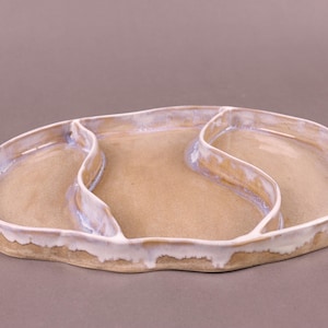 Könnte beinhalten: Eine Servierplatte aus Keramik mit drei Abteilen. Die Platte ist hellbraun mit einer weißen Glasur. Die Platte hat eine organische, freiförmige Form.