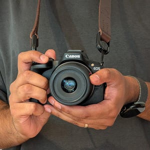May include: A black Canon EOS camera with a lens, held by a person. The camera has a brown leather strap. The person is wearing a grey shirt and a black and grey watch.