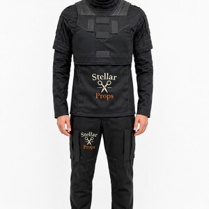 May include: A black tactical-style uniform consisting of a long-sleeved shirt with a high collar, a chest plate overlay, and matching trousers. The shirt and pants feature the text Stellar Props with a scissors icon printed in white and orange.