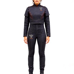 May include: A dark charcoal gray jumpsuit featuring a high-neck cropped jacket overlay with vertical stitching and long sleeves. The garment includes a logo with the text Stellar Props on the chest and thigh, paired with matching dark boots.