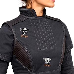 May include: A black high-neck tunic featuring quilted shoulder and side panels with visible stitching. The garment displays the text Stellar Props accompanied by a white scissors icon printed twice on the front chest area in a light color.