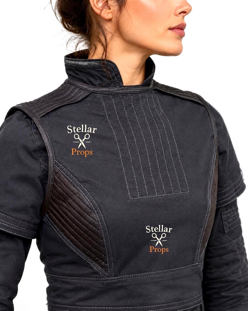 May include: A black high-neck tunic featuring quilted shoulder and side panels with visible stitching. The garment displays the text Stellar Props accompanied by a white scissors icon printed twice on the front chest area in a light color.