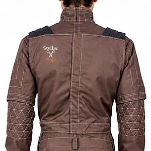 May include: A brown jacket with a high collar, featuring white contrast stitching, black shoulder accents, and quilted forearm panels. The back displays the text Stellar Props with a scissors icon embroidered in white and orange thread.