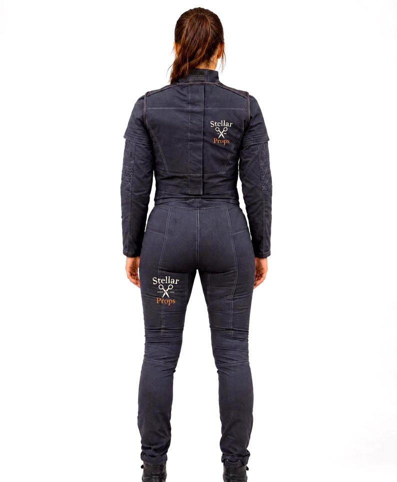 May include: A dark gray jumpsuit featuring long sleeves, a high collar, and visible seam detailing. The back of the jacket and the back of the right leg display the white and orange text Stellar Props with a graphic icon of crossed scissors.