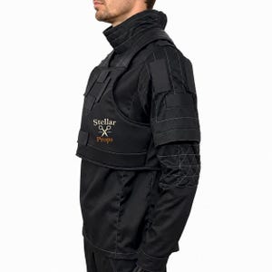May include: A black tactical-style vest worn over a matching black long-sleeve shirt. The vest features a high collar, structured panels, and the text Stellar Props with a scissors icon printed in white and orange on the chest area.