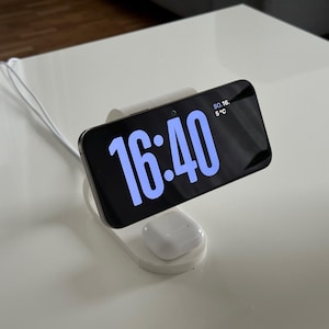 May include: A white charging station with a black smartphone displaying the time 16:40 in blue. A pair of white wireless earbuds rests on the base. The phone shows the date and temperature.