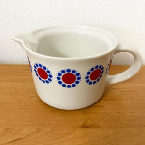 Op de afbeelding: Witte keramische creamer met een handvat en tuit. De creamer is versierd met een herhalend patroon van rode en blauwe bloemmotieven. De creamer staat op een houten oppervlak.