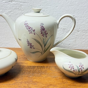 Könnte beinhalten: Ein cremefarbenes Teeservice mit Teekanne, Zuckerdose und Sahnekännchen. Das Set ist mit lila Lavendelzweigen und grünen Blättern verziert. Zarte Libellenillustrationen sind ebenfalls auf Zuckerdose und Teekanne vorhanden.