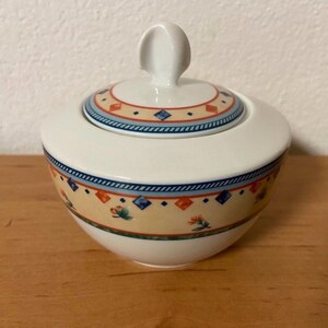 Könnte beinhalten: Weiße Porzellan-Zuckerdose mit Deckel. Die Schale hat einen blauen, orangen und gelben floralen Muster um den Rand.
