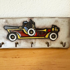 Könnte beinhalten: Ein rechteckiger Schlüsselhalter mit einem Oldtimer-Design. Das Auto ist hauptsächlich rot mit gelben und schwarzen Akzenten und einer Fahrerfigur. Der Halter hat fünf Metallhaken für Schlüssel und einen verwitterten, cremefarbenen Hintergrund.