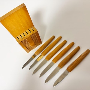 Könnte beinhalten: Ein Messerblock aus Holz mit sechs Messern. Die Messer haben Holzgriffe und silberne Klingen. Der Messerblock ist aus hellem Holz gefertigt und hat Schlitze für die Messer.