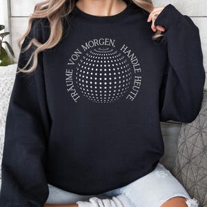 Könnte beinhalten: Schwarzer Pullover mit einem weißen Grafikdesign einer Kugel mit dem Text "Träume von Morgen, Handle Heute" darum herum.