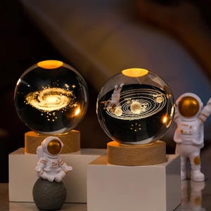 May include: Two glass globes with a wooden base, one depicting a galaxy and the other depicting the solar system.  Each globe is illuminated from within, creating a warm glow.  A small astronaut figurine sits on a moon-shaped base in front of the galaxy globe.