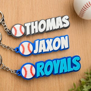 Könnte beinhalten: Drei personalisierte Schlüsselanhänger mit einem weißen Baseball-Symbol mit roten Nähten, befestigt an einem Metallring. Die Anhänger zeigen die Namen THOMAS in Weiß auf schwarzer Basis, JAXON in Weiß auf blauer Basis und ROYALS in Blau auf hellblauer Basis.