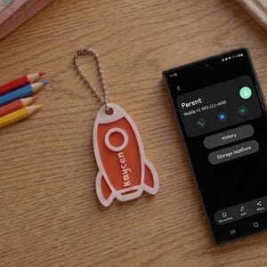 Pode incluir: Um porta-chaves em forma de foguetão branco e laranja com uma corrente de esferas prateada, com o nome Kaycen impresso verticalmente. O porta-chaves está colocado ao lado de um smartphone que mostra um ecrã de contacto Parent, um estojo às riscas e um caderno.