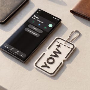 Pode incluir: Um smartphone preto exibindo uma tela de contato, um porta-passaporte escuro, óculos de sol com armação marrom, uma carteira de couro marrom e uma etiqueta de bagagem retangular branca com borda preta e o texto YOW com ícone de avião.