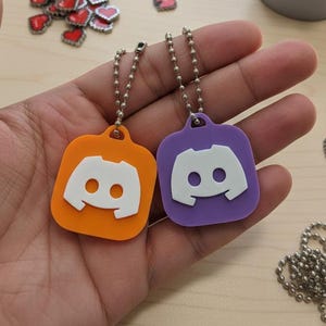 May include: Two keychains with the Discord logo, one orange and one purple, are held in a hand. Each keychain has a white logo and a silver chain. Small heart-shaped decorations are scattered around the keychains.
