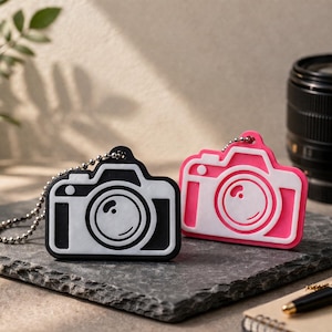 Può includere: Due portachiavi a forma di fotocamera, uno nero e uno rosa, con dettagli bianchi. Ogni portachiavi ha una catena d'argento. Un obiettivo della fotocamera e una penna sono visibili sullo sfondo. I portachiavi sono su una superficie di pietra grigio scuro.