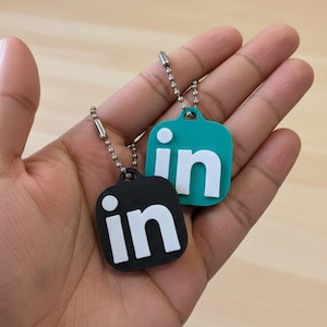 Może przedstawiać: Dwa breloki do kluczy z logo LinkedIn. Jeden brelok jest czarny z białymi literami, drugi turkusowy z białymi literami. Każdy brelok ma srebrny łańcuszek.