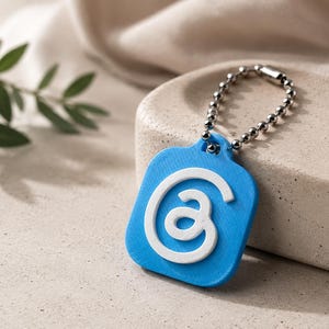 Può includere: Un portachiavi quadrato blu brillante con angoli arrotondati caratterizzato da un simbolo a spirale bianco in rilievo al centro. Il portachiavi è fissato a una catenella a palline in metallo argentato, creando un accessorio funzionale e contrastato.