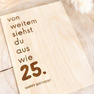 Könnte beinhalten: Eine hellfarbene Holz-Geburtstagskarte mit dem eingravierten Text "von weitem siehst du aus wie 25. HAPPY BIRTHDAY." Die Karte ist rechteckig und liegt auf einer hellfarbenen Holzoberfläche.