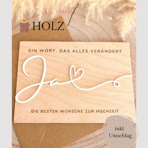 Könnte beinhalten: Eine Holz-Hochzeitskarte mit dem Wort "Ja" in Schreibschrift, begleitet von dem Text "EIN WORT, DAS ALLES VERÄNDERT" und "DIE BESTEN WÜNSCHE ZUR HOCHZEIT". Die Karte liegt auf einem braunen Umschlag, mit dem Wort "HOLZ" oben.
