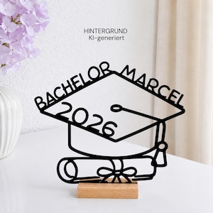 Könnte beinhalten: Eine schwarze Metall-Silhouette einer Doktorhut-Form mit dem Text BACHELOR MARCEL und 2026 sowie einem Diplom-Symbol an der Basis, montiert auf einem rechteckigen hellen Holzsockel. Das Design zeigt eine Quaste an der rechten Seite des Hutes.