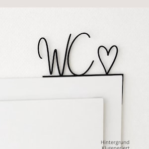 Könnte beinhalten: Ein Türschild aus schwarzem Metall mit den Buchstaben WC in Schreibschrift, gefolgt von einem Herzumriss. Das Schild hat eine flache horizontale Basis, die auf der Oberkante des Türrahmens aufliegt, und eine vertikale Lasche zur Befestigung.