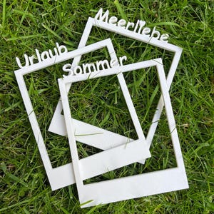 Könnte beinhalten: Drei weiße, rechteckige Fotorahmen im klassischen Sofortbildkamera-Design. Jeder Rahmen trägt oben ein Wort: Meerliebe, Urlaub und Sommer. Die Rahmen sind übereinander gestapelt und bilden eine geschichtete Anordnung aus weißen Rändern.