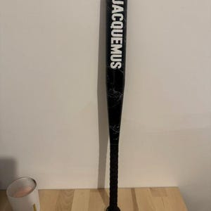 Puede incluir: Un bate de béisbol negro con un patrón de mármol blanco y la palabra JACQUEMUS impresa en letras mayúsculas blancas grandes verticalmente a lo largo del cuerpo. El mango tiene un agarre negro texturizado y un pomo redondeado en la base.