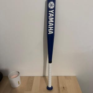 Op de afbeelding: Een blauwe honkbalknuppel met het witte Yamaha-logo en de tekst verticaal op de loop gedrukt. Het handvat is omwikkeld met wit gripband en de knuppel eindigt in een blauwe knop. Het ontwerp is strak, opvallend en toont duidelijk de merknaam.
