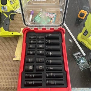Puede incluir: Maleta Milwaukee Packout roja abierta con tapa transparente, que contiene un juego de vasos de impacto negros. La maleta está sobre una mesa con otras herramientas, incluido un taladro amarillo Ryobi.