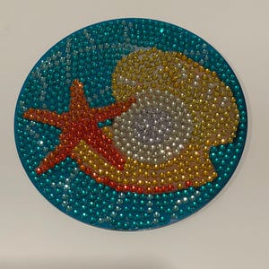 May include: A round, blue, sparkly, resin-based, decorative piece with a seashell and starfish design. The seashell is yellow, orange, and white. The starfish is orange and red.
