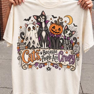 May include: A cream-coloured t-shirt featuring a graphic of four cats in Halloween costumes, including a ghost, a vampire, a pumpkin-headed cat, and a skeleton cat. The text reads, "Cats, Because People Are Creepy" and "Creep It Cute" on a tombstone.