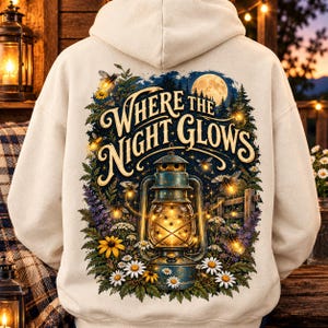 Puede incluir: Una sudadera con capucha de color crema que presenta un gráfico en la espalda de una linterna brillante rodeada de flores silvestres, luciérnagas y una luna llena. El texto "WHERE THE NIGHT GLOWS" aparece en una fuente decorativa.