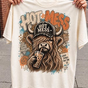 May include: An off-white t-shirt featuring a graphic of a Highland cow wearing a black baseball cap labeled HOT MESS. The cow drinks bubble tea. Text includes HOT MESS, BUT STILL CUTE, ICED COFFEE BIGGER PROBLEMS, and GOOD THINGS TAKE TIME.